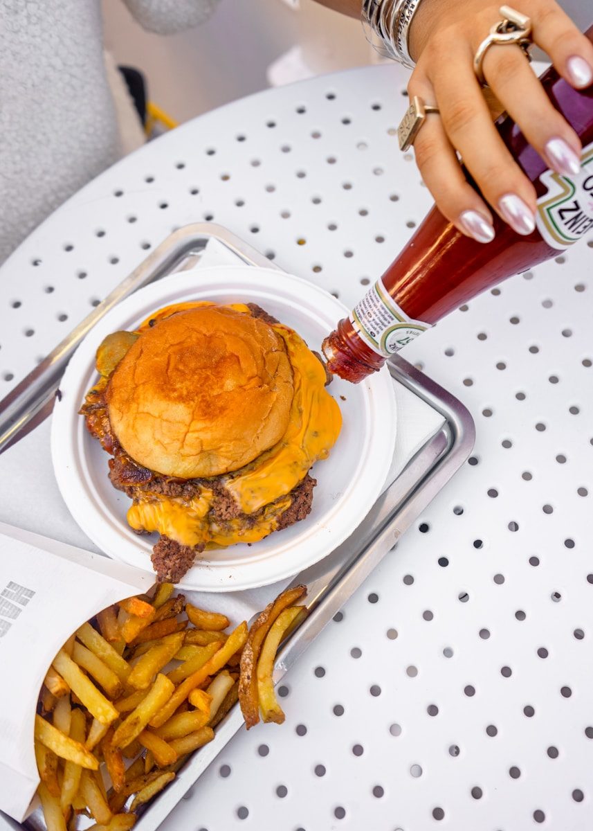 Hand adding ketchup to a double cheeseburger and fries.
