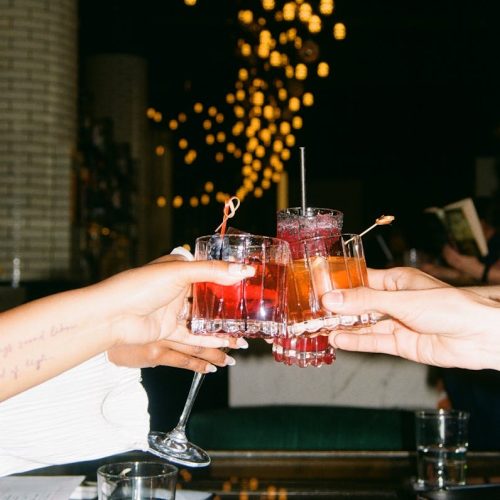Friends raise colorful cocktails in a vibrant bar setting, celebrating casually.